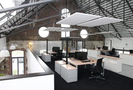 Ein modernes Büro im Obergeschoss eines Industriegebäudes mit hohen Decken und großen Fenstern. Mehrere Schreibtische mit Computern und schwarzen Bürostühlen sind im Raum verteilt, während schallabsorbierende Paneele von der Decke hängen.