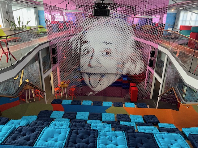 Empore mit Sitzgelegenheiten hinter einem Raumtrenner mit Einstein-Gesicht, auf der Treppe Sitzkissen