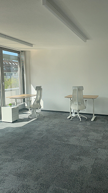 Büroraum mit zwei Schreibtischen, weißen Bürostühlen und grauem Teppichboden