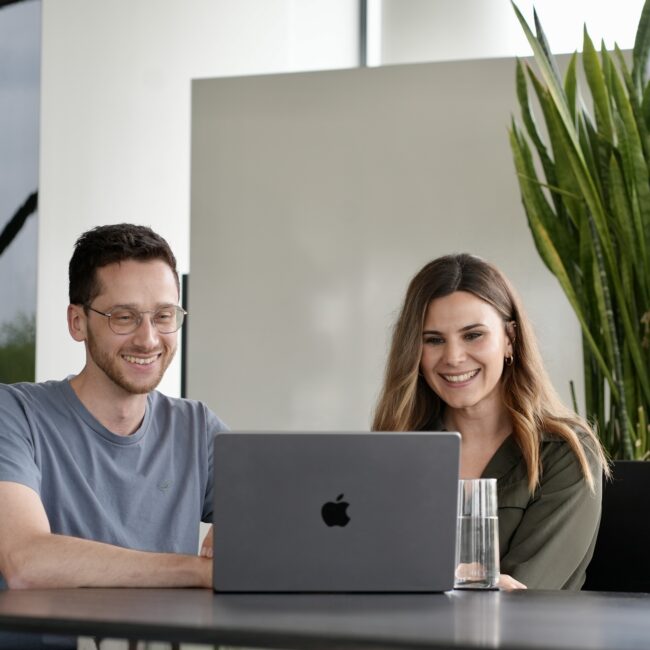 Zwei Personen sitzen an einem Tisch, schauen auf einen Laptop und lächeln.
