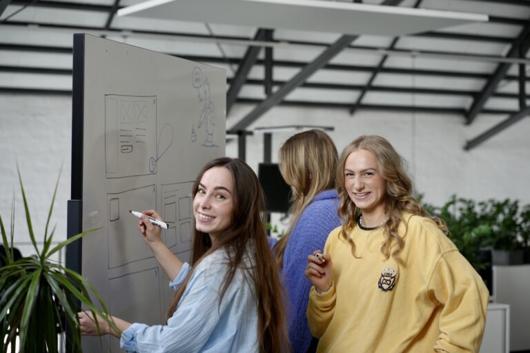 Drei Personen stehen lächelnd an einem Whiteboard und bemalen dieses.