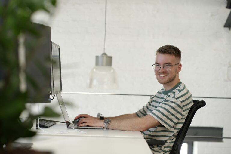 Ein Mann sitzt an einem Schreibtisch, arbeitet an einem Laptop und lacht in die Kamera.