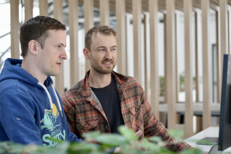 Zwei Männer sitzen nebeneinander vor einem Schreibtisch und schauen auf einen Bildschirm.