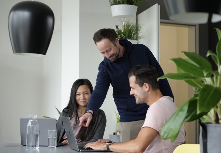 Zwei Personen sitzen an einem Tisch und schauen auf Laptops, wobei eine weitere Person daneben steht und auf einen dieser Laptops zeigt.