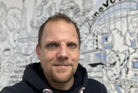 Nils Domrose mit Hoodie steht lächelnd vor einem inovex-Comic-Mural
