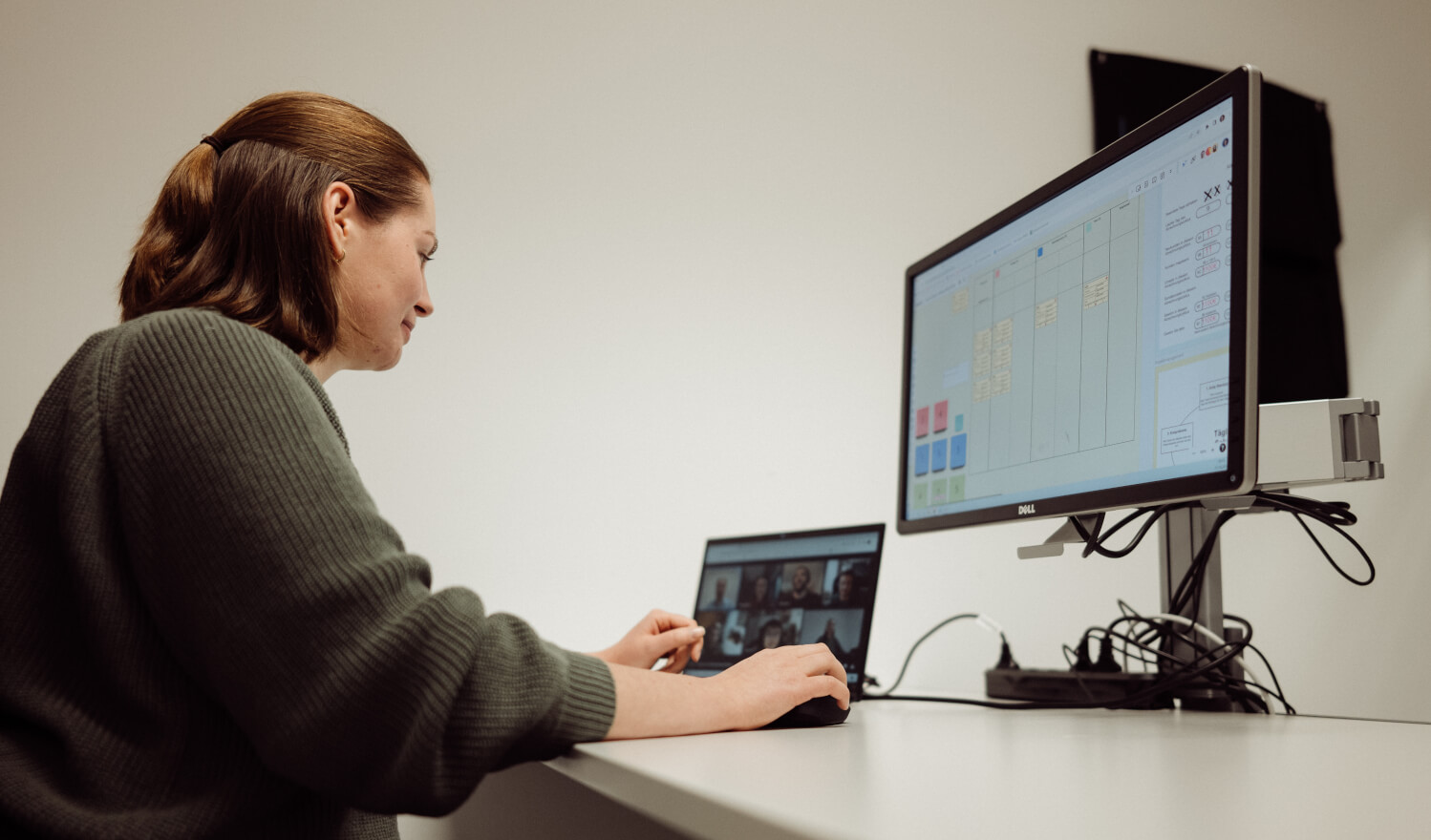 Eine Person sitzt lächelnd vor einem Laptop mit virtuellen Meeting und einem zweiten Bildschirm mit einem Board