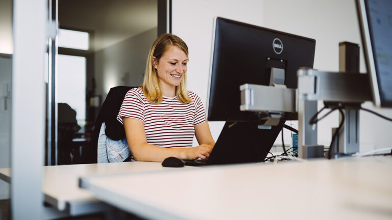 Eine lächelnde Person sitzt am Schreibtisch mit Bildschirm und Laptop