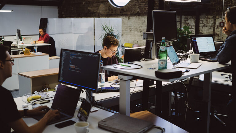 Vier Personen sitzen oder stehen verteilt in einem Büro an Schreibtischen mit Bildschirmen und Laptops