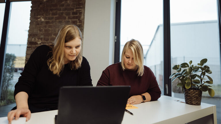 Zwei Personen sitzen an einem Tisch, eine schreibt, die andere schaut auf einen Laptop