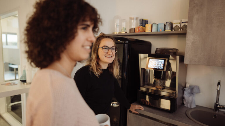 Zwei Personen stehend lächelnd in einer Küche vor Kühlschrank und Kaffeemaschine, eine hält eine Tasse