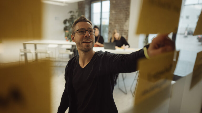 Person hinter einer Glaswand zeigt auf ein beschriftetes PostIt an der Glaswand