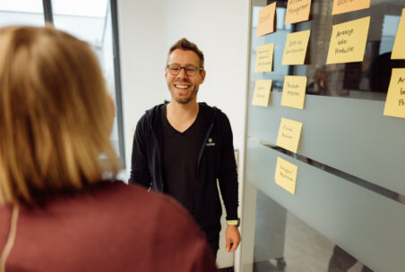 Zwei lächelnde Personen stehen neben einer Glaswand, auf die gelbe und orangene Post-Its geklebt wurden