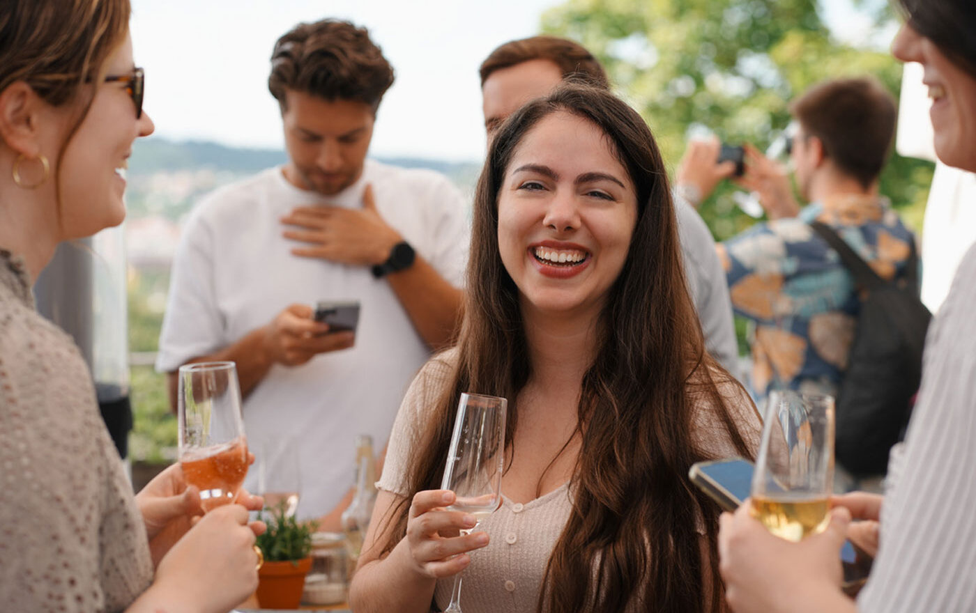 Mehrere Menschen stehen mit Gläser in der Hand draußen in einer Gruppe. Eine Person lächelt direkt in die Kamera. Im Hintergrund sind ebenfalls Menschen.