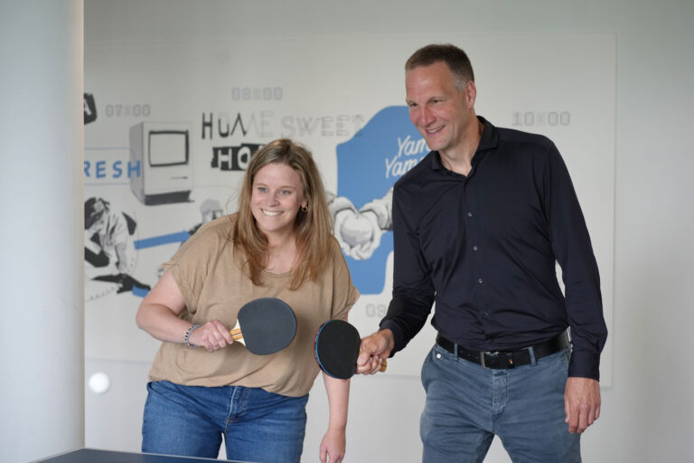 Zwei Personen haben Tischtennisschläger in den Händen und stehen nebeneinander vor einer Tischtennisplatte.