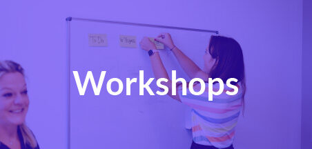 Das Wort Workshops auf blau gefärbtem Foto von einer Frau, die eine Haftnotiz an ein Whiteboard klebt. Eine zweite Frau blickt in die Kamera.